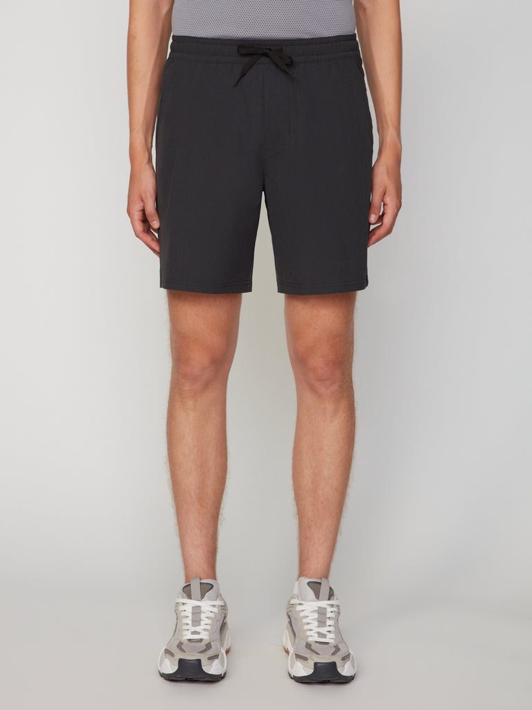 A person, visible from the waist down against a plain light background, wears a gray shirt and TECH APPAREL MEN’S 360:FLEX PULL-ON SHORT – 7.5 INSEAM in black, paired with white sneakers.