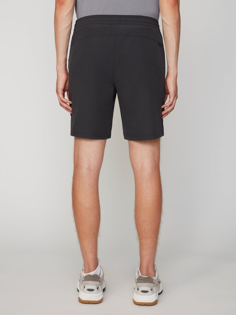 A person faces away from the camera wearing TECH APPAREL MEN’S 360:FLEX PULL-ON SHORT – 7.5 INSEAM in black, paired with a grey shirt and white sneakers, set against a plain light background.
