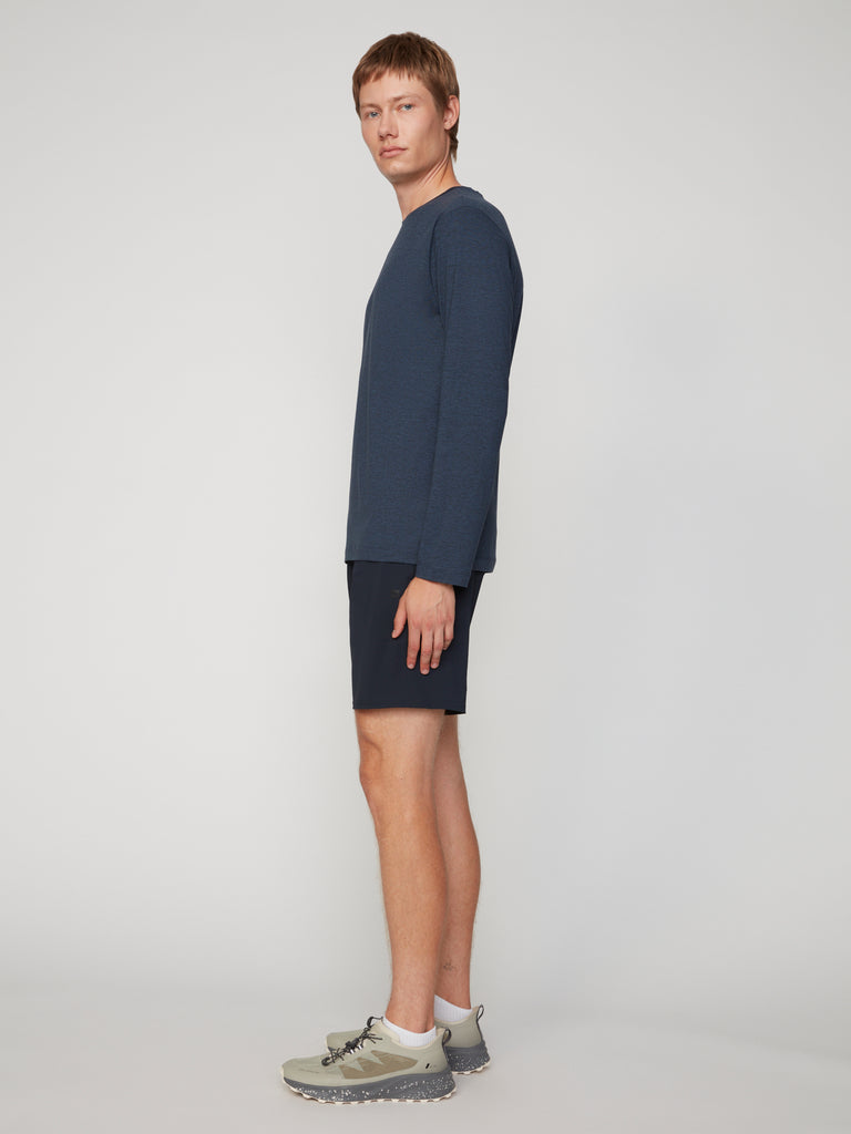 A man faces sideways toward the camera, wearing a long-sleeve dark blue shirt, TECH APPAREL MEN’S FLEXLITE PULL-ON SHORT (8 inseam) made from stretch FlexLite fabric, and gray sneakers, with a plain light background.