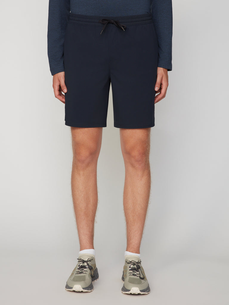 A person wears TECH APPAREL MEN’S FLEXLITE PULL-ON SHORT, 8 INSEAM in navy blue with light green athletic shoes, standing against a plain light gray background. Only the lower body is visible.
