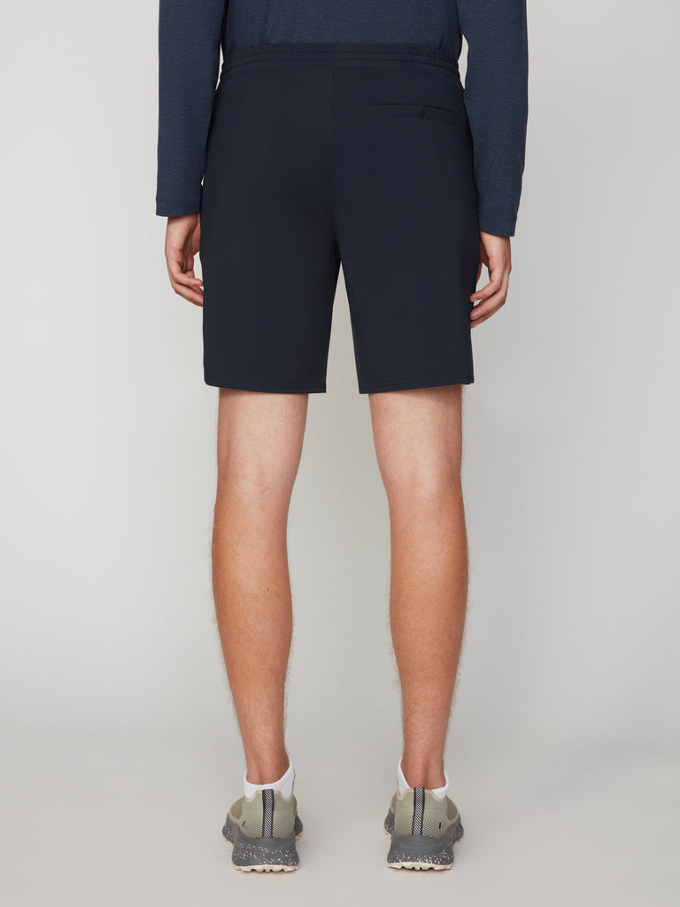 A person wearing a long-sleeve navy shirt, TECH APPAREL MEN’S FLEXLITE PULL-ON SHORT, 8 INSEAM, and gray athletic shoes stands facing away from the camera against a plain light background.