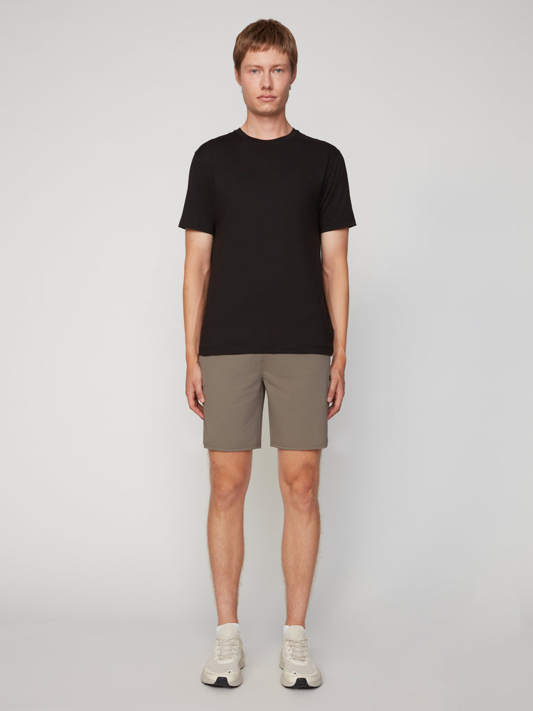 A man stands facing forward against a plain light gray background, wearing a black t-shirt, white sneakers, and TECH APPAREL MEN’S FLEXLITE PULL-ON SHORT, 8 INSEAM. His arms rest naturally at his sides.