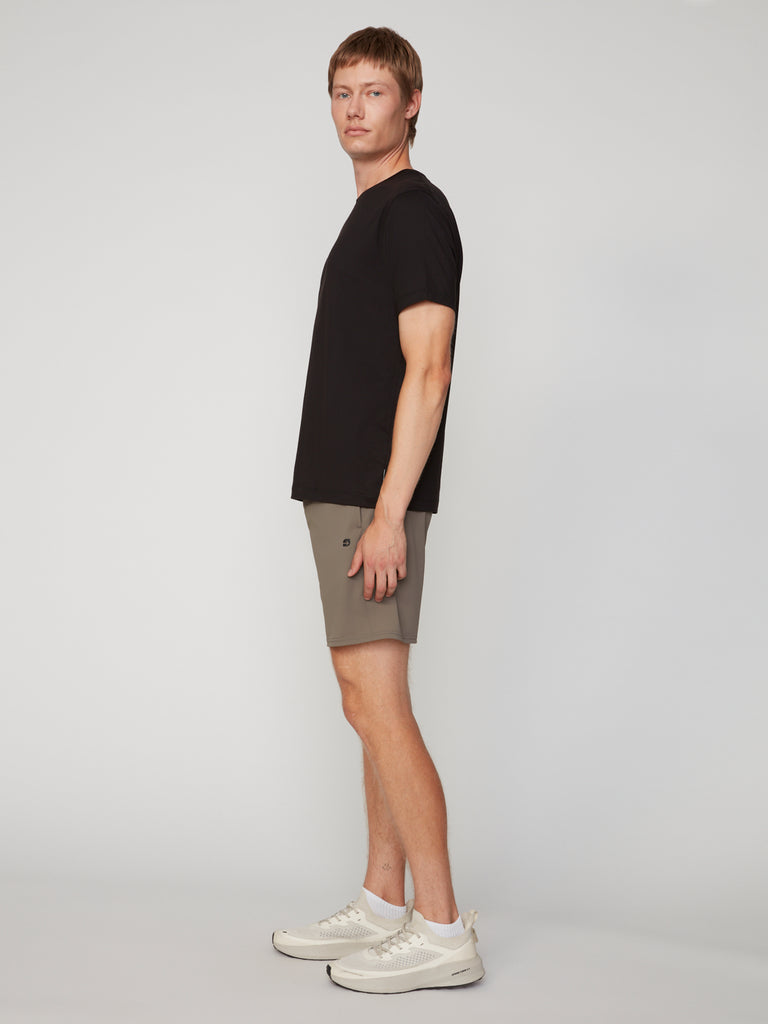 A man stands sideways against a plain background, wearing TECH APPAREL’s MEN’S FLEXLITE PULL-ON SHORT (8 inseam) with a black t-shirt and white sneakers, looking straight ahead with a neutral expression.