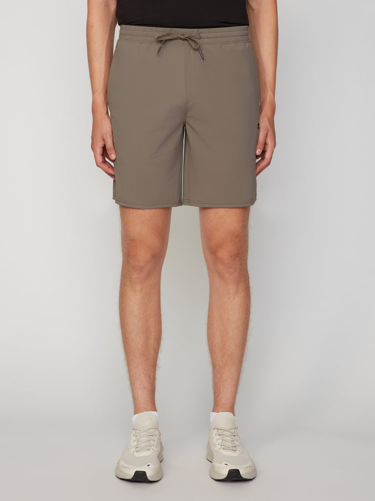 A man stands against a plain background, wearing TECH APPAREL MEN’S FLEXLITE PULL-ON SHORT (8 inseam) in taupe, styled with a black top and white sneakers.