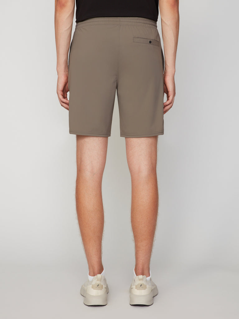 A person stands facing away, wearing TECH APPAREL MEN’S FLEXLITE PULL-ON SHORT (8 inseam) in taupe, featuring lightweight FlexLite fabric, an elastic waistband, a back pocket, light sneakers, and a black shirt.